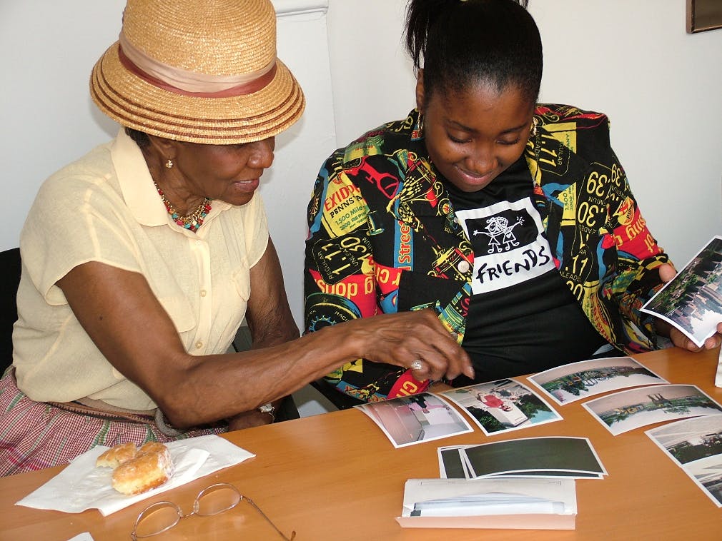 Two Black women sit side by side at a table looking at photographs together. The women on the left wears a yellow shirt and straw brimmed hat. The woman on the right wears her hair pulled back, a floral button down shirt open, and a black t-shirt underneath.