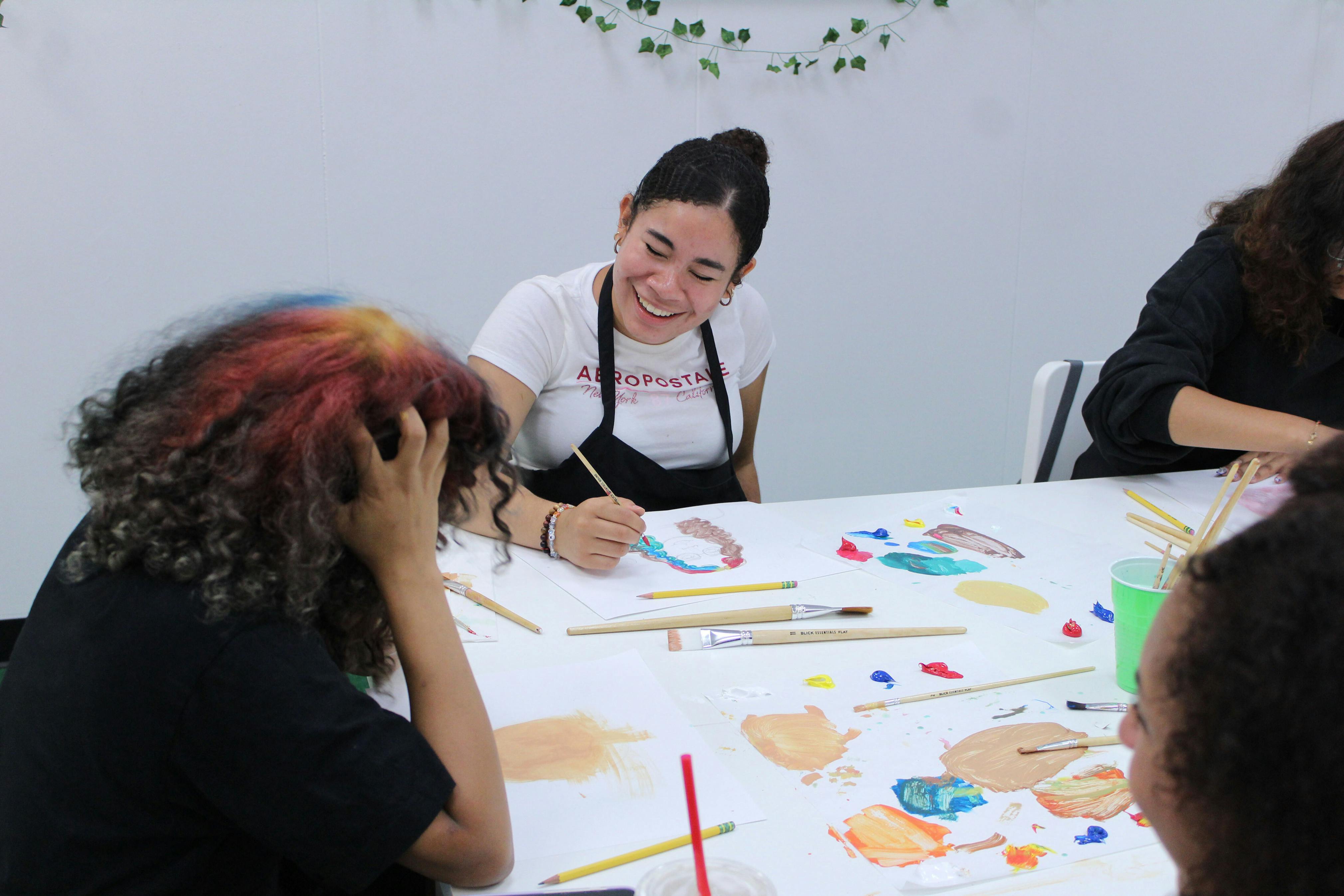 Teens work on an activity at a white table and the teen facing the camera is smiling with her head tilted.