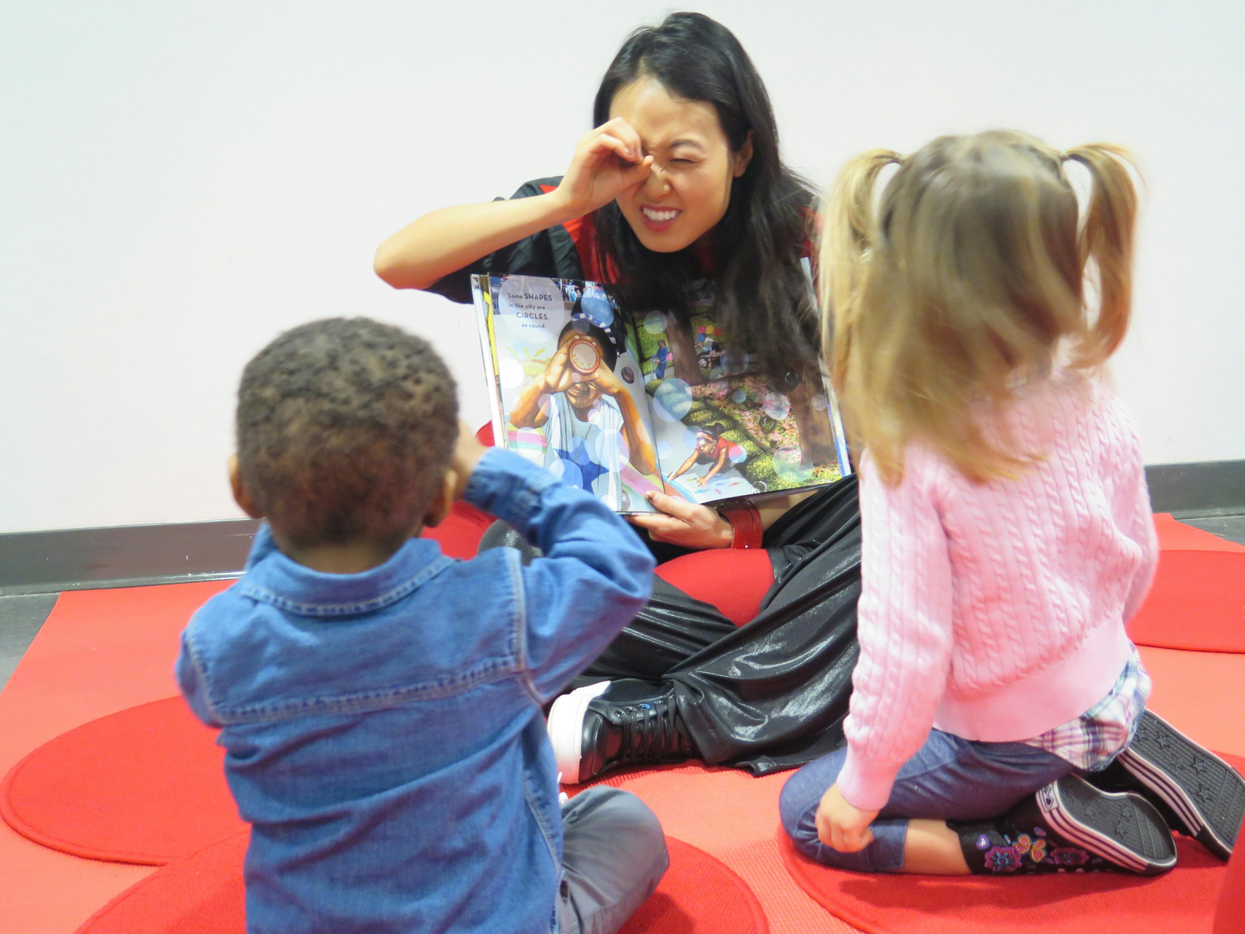 Two children face a museum-educator reading them a story on a red rug.