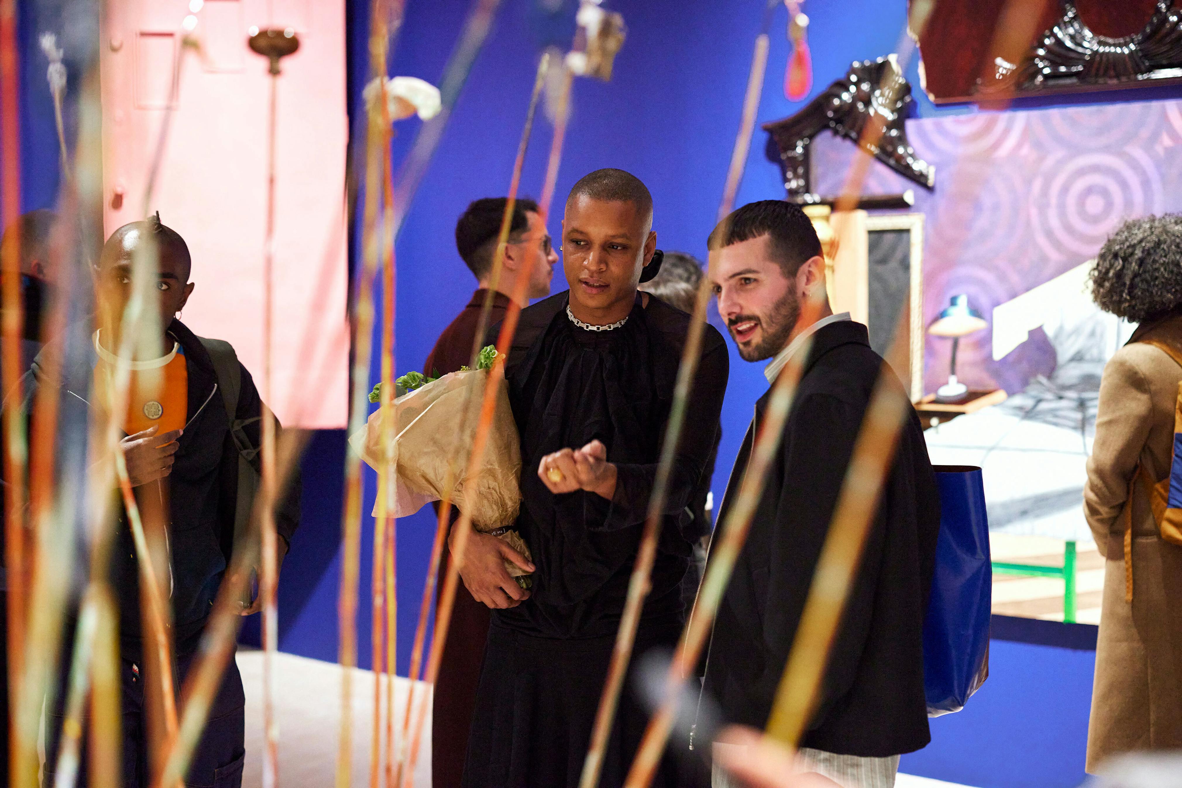 Two people wearing black stand in front of artwork on a blue wall looking at and through standing stick-like objects.