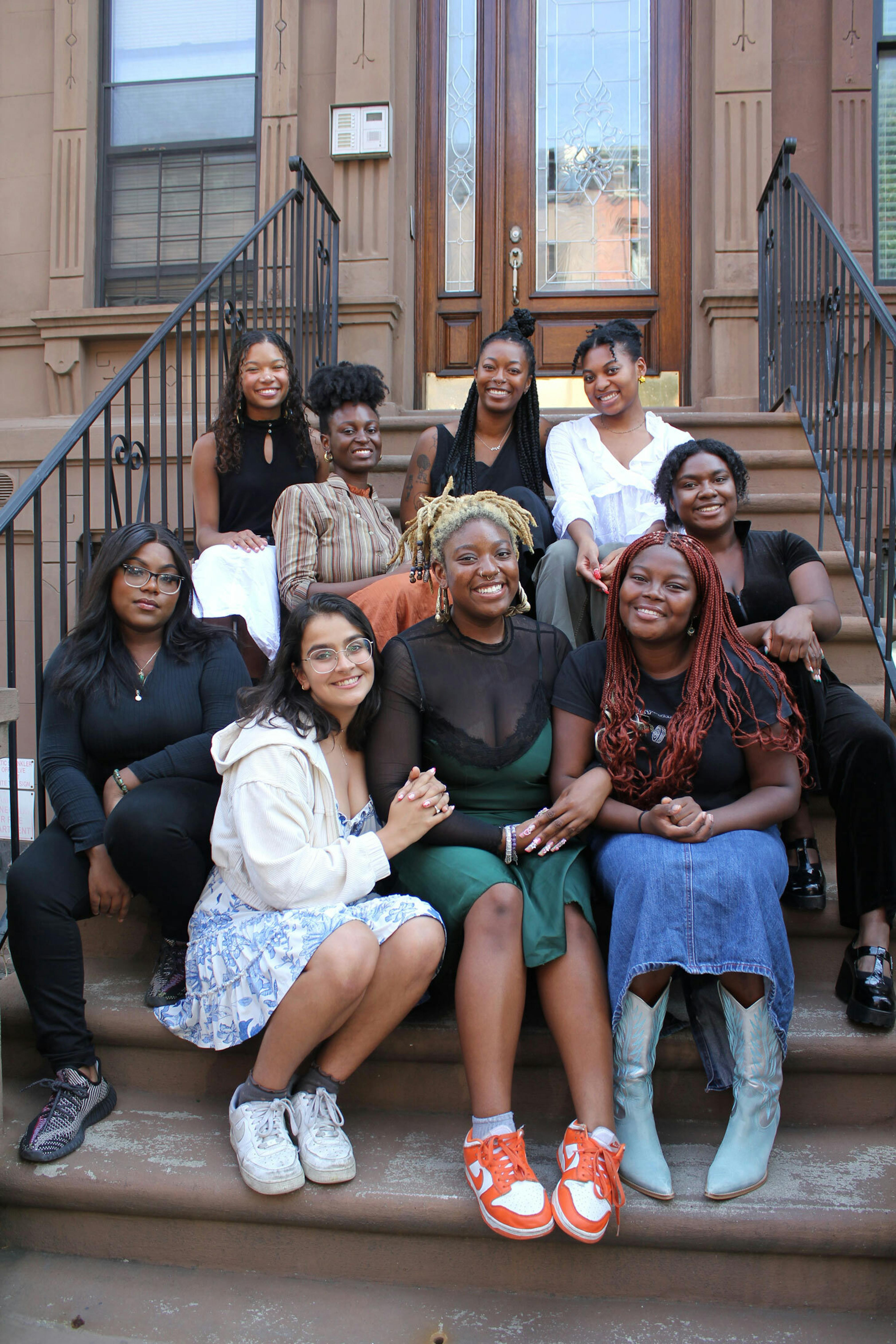 A group of smiling young adults sit together on the stoop of a brownstone.