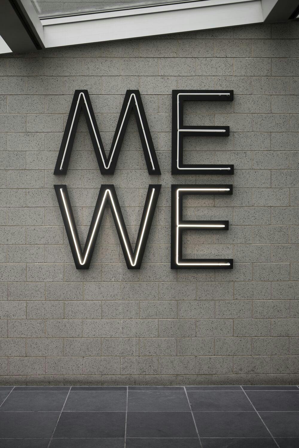 A neon sculpture with the word Me stacked on top of the word We is hung on a grey wall.