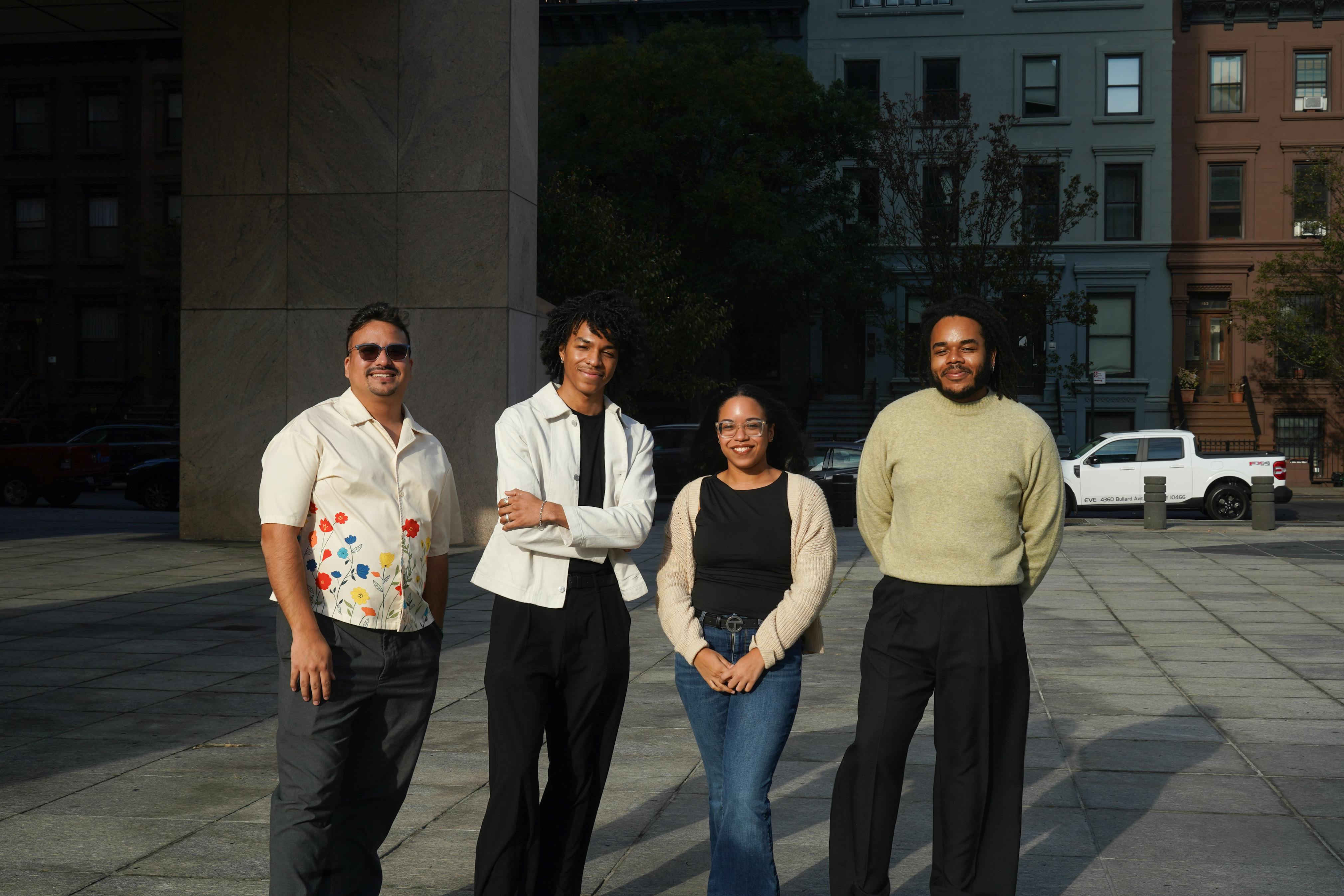 Three brown skinned men and one brown skinned woman stand next to each other and smile at the camera. A white car and apartment buildings can be seen in the background