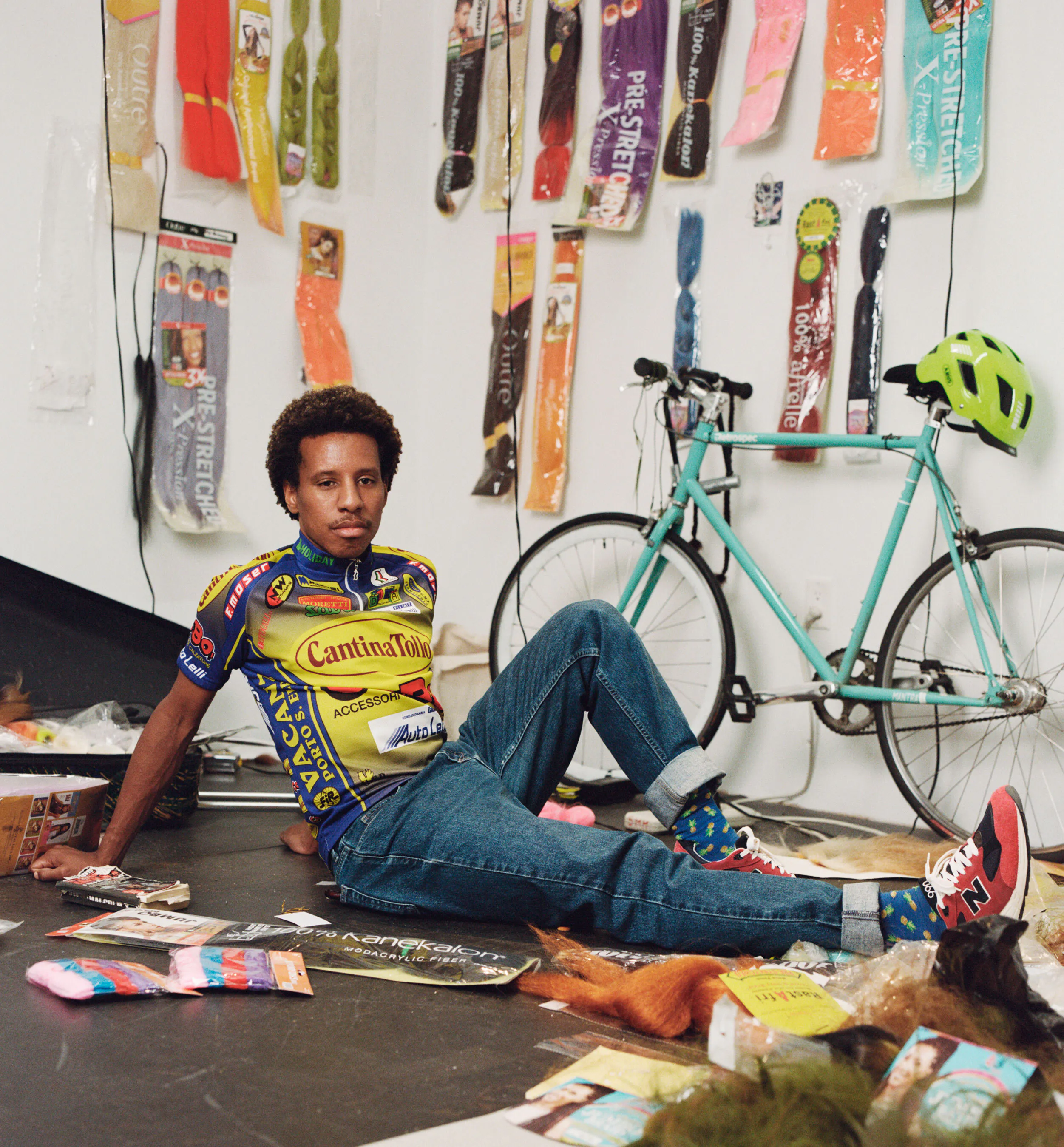 A brown-skinned person lounges on the floor of a cluttered studio.