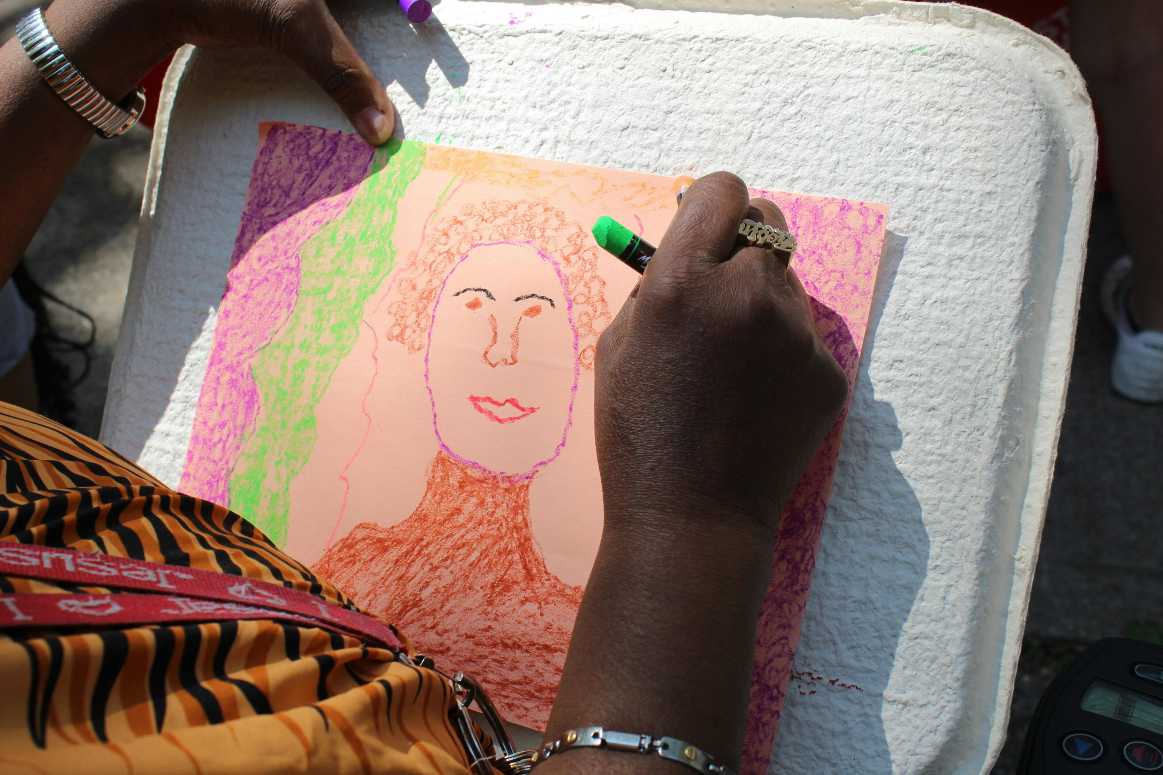 Close up of dark brown hand holding a green colored pastel to draw a portrait on tan paper.